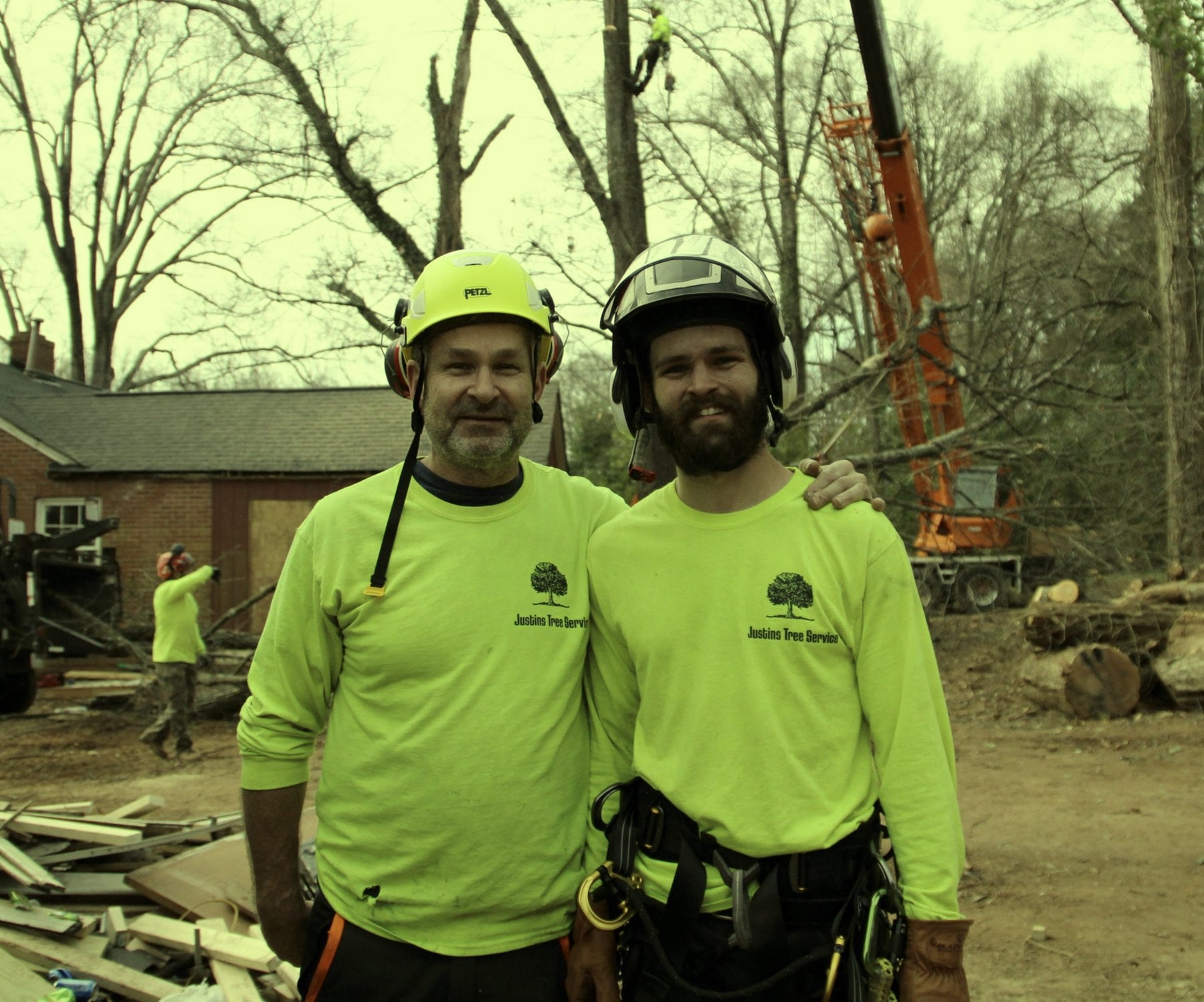 Two Justin's Tree Service crew members in hi-vis lime company shirts and helmets, arms around each other, with a climber up the tree and an orange crane behind them